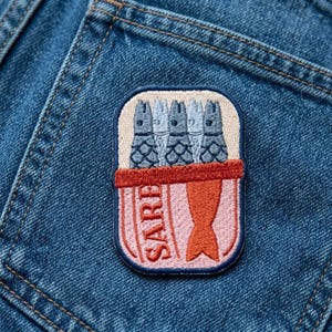 May include: An embroidered patch featuring a rectangular tin of sardines with a dark blue border. The design shows four grey fish heads and one orange fish tail, with the text SARDINES printed vertically in orange on a light pink background.