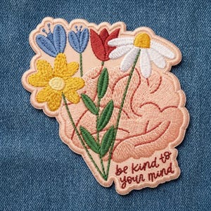May include: An embroidered patch featuring a peach-colored brain shape with a bouquet of flowers including a yellow daisy, two blue bell-shaped flowers, a red tulip, and a white daisy. The text "be kind to your mind" is stitched in brown.