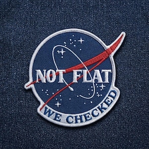 May include: A circular embroidered patch featuring a dark blue background with white stars and a white orbital line. A red swoosh crosses the center. The text NOT FLAT is in white, with WE CHECKED in white on a curved banner at the bottom edge.