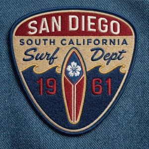 May include: A shield-shaped embroidered patch on blue denim fabric featuring the text SAN DIEGO SOUTH CALIFORNIA Surf Dept 1961. The design includes a central red and white surfboard with a white flower icon, set against dark blue waves.
