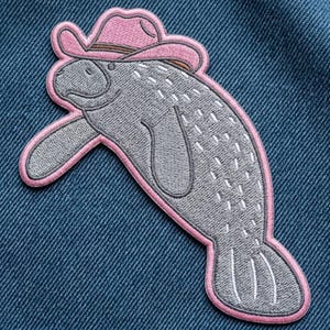 May include: An embroidered patch featuring a gray manatee wearing a pink cowboy hat with a brown band. The manatee has white stitched details on its body and a pink border outline, all set against a dark blue denim fabric background.