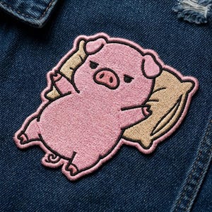 May include: An embroidered patch featuring a pink pig lying on a tan pillow. The pig has a rounded body, small ears, and a curled tail, with a neutral expression. The patch is outlined in a thick pink border and is attached to dark blue denim fabric.