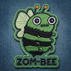 May include: An embroidered patch featuring a green and black striped bee character with large white eyes, a stitched mouth, and light green wings. Two antennae with orange tips extend from the top. The text ZOM-BEE is embroidered in black at the base.