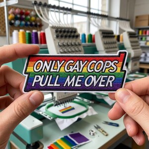 May include: A rectangular embroidered patch featuring a rainbow-colored background with the white text "ONLY GAY COPS PULL ME OVER" in a bold, sans-serif font. The patch has a thick white border and is held against a blurred industrial background.
