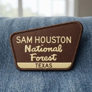 May include: A brown embroidered patch with a trapezoidal shape featuring the text SAM HOUSTON National Forest in cream-colored lettering, with the word TEXAS printed in brown on a cream-colored horizontal bar at the bottom of the patch.