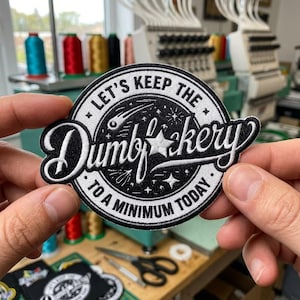 May include: A circular embroidered patch featuring a black background with white stitching. The text reads "LET'S KEEP THE DUMB-KERY TO A MINIMUM TODAY" in a stylized font, accented by a shooting star, a five-pointed star, and small sparkles.
