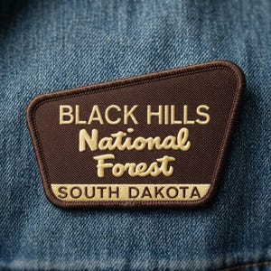 May include: A brown embroidered patch with a dark brown border featuring the text BLACK HILLS National Forest in cream-colored lettering, with the text SOUTH DAKOTA in a cream-colored bar at the bottom, all set against a blue denim fabric background.