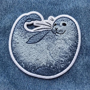 May include: A circular embroidered patch featuring a gray seal with detailed stitching, dark eyes, and whiskers. The seal is curled into a rounded shape, showcasing textured fur patterns in shades of gray and white, finished with a white border.