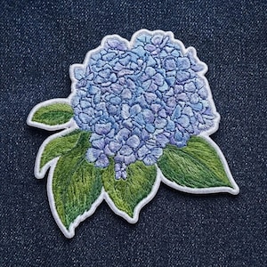 May include: An embroidered patch featuring a cluster of light blue and periwinkle hydrangea flowers with detailed green leaves. The design has a white stitched border and is shaped to follow the contours of the floral and leaf arrangement.