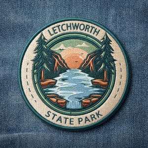 May include: A circular embroidered patch featuring a landscape scene of Letchworth State Park. The design includes a central waterfall, dark green pine trees, rocky terrain, and an orange sun against a light background with the text Letchworth State Park.