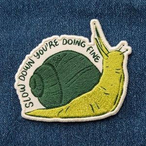 May include: An embroidered patch featuring a yellow-green snail with a dark green shell. The text 'SLOW DOWN YOU'RE DOING FINE' is stitched in dark green letters along the cream-colored border that curves around the snail's shell.