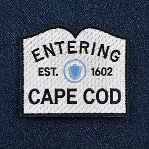 May include: A white embroidered patch with a black border and black text that reads ENTERING EST. 1602 CAPE COD. The patch features a central light blue circular emblem containing a shield icon, all set against a dark blue denim fabric background.
