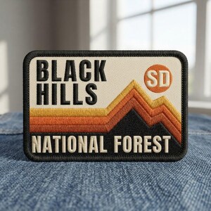 May include: A rectangular embroidered patch featuring the text BLACK HILLS NATIONAL FOREST in black and cream lettering. The design includes a mountain range graphic in yellow, orange, and black, with a circular orange emblem containing the letters SD.