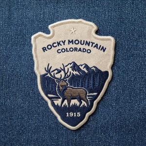 May include: A shield-shaped embroidered patch featuring a cream-colored background with dark blue text reading ROCKY MOUNTAIN COLORADO and 1915. The design includes a dark blue mountain range, pine trees, and a brown elk with large antlers.