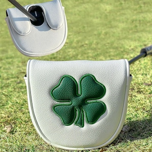 May include: A white textured golf putter headcover featuring a large green four-leaf clover appliqué on the front. The cover has a rounded shape with a black interior lining and a small opening at the top for the putter shaft to pass through.
