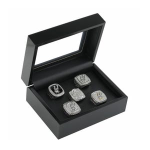 May include: A black wooden display box with a clear hinged lid containing five silver-toned championship rings. Each ring features intricate detailing, embedded clear stones, and the word CHAMPIONS engraved on the metal surface of the bands.