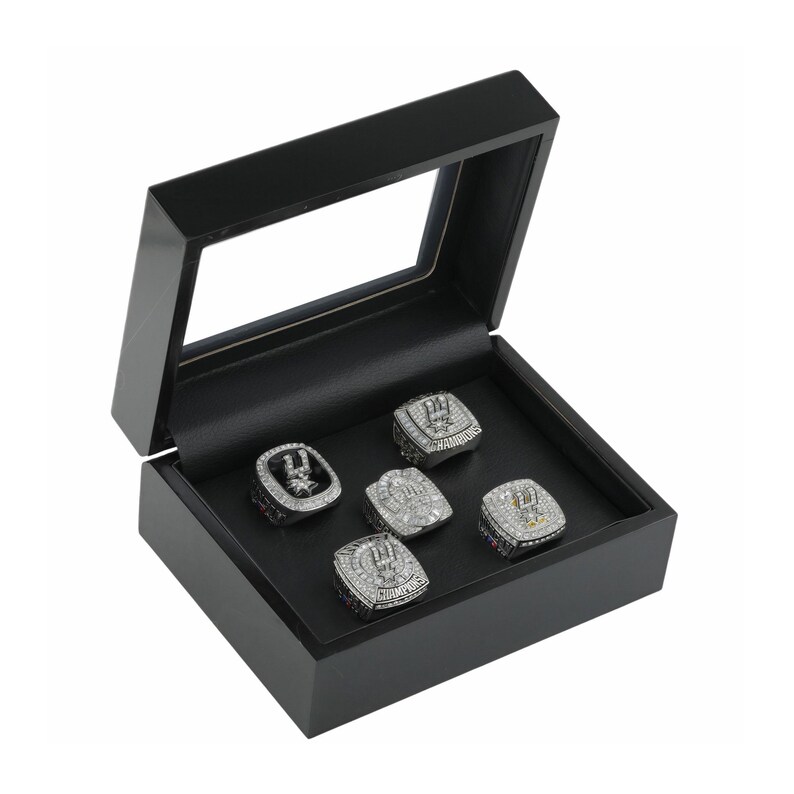 May include: A black wooden display box with a clear hinged lid containing five silver-toned championship rings. Each ring features intricate detailing, embedded clear stones, and the word CHAMPIONS engraved on the metal surface of the bands.