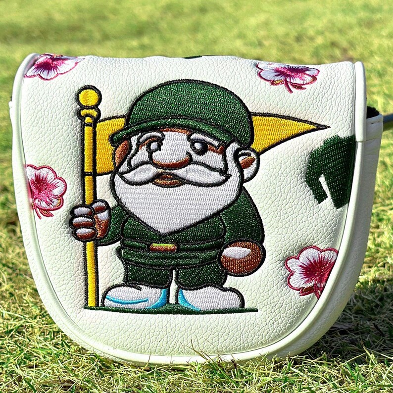 May include: A white golf putter headcover featuring an embroidered gnome wearing a green hat and outfit, holding a yellow flag. The cover is decorated with pink and white floral patterns across the textured white surface of the accessory.