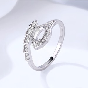 Puede incluir: Un anillo plateado con un diseño único. El anillo presenta una forma ovalada central y un elemento lateral decorativo, ambos incrustados con pequeñas piedras preciosas transparentes. El anillo se encuentra sobre un fondo suave y claro, resaltando sus detalles.