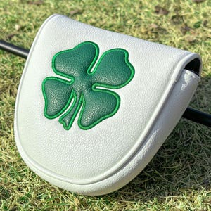 May include: A white golf putter headcover featuring a textured surface and a large, dark green four-leaf clover appliqu&eacute; centered on the front. The cover has a rounded shape with white piping along the edges and a black shaft visible behind it.