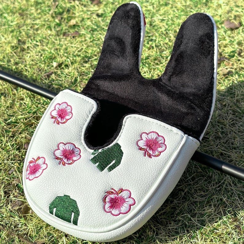 May include: A white mallet-style golf putter headcover featuring a black interior lining. The exterior is decorated with embroidered pink flower motifs and small green golf sweater icons scattered across the textured white surface.