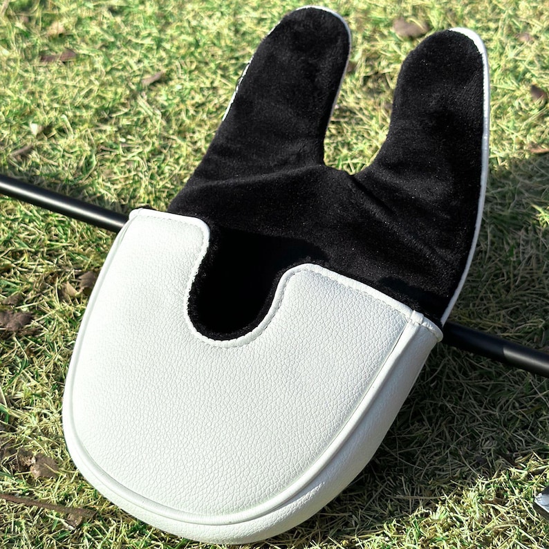 May include: A mallet-style golf putter headcover featuring a white textured exterior and a soft black interior lining. The cover has a U-shaped opening designed to fit over the putter head, with a smooth, rounded front edge and a two-pronged top.
