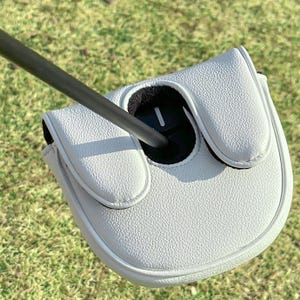 May include: A white textured golf putter headcover with a black interior lining and a single white vertical line detail. The headcover is attached to a thin black shaft that extends diagonally across the frame, showcasing a clean design.