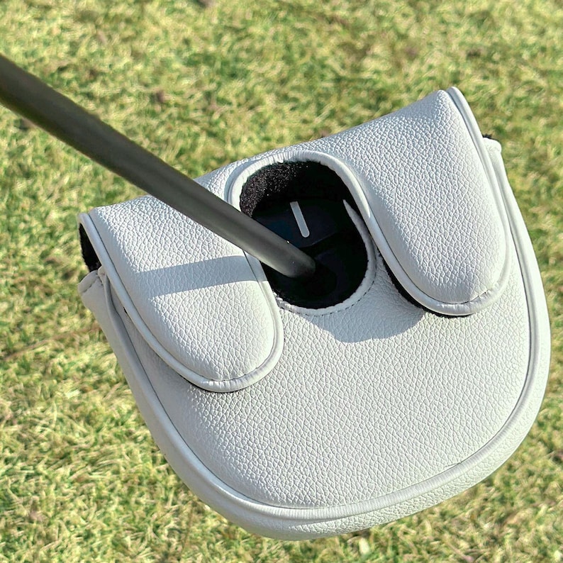 May include: A white textured golf putter headcover with a black interior lining and a single white vertical line detail. The headcover is attached to a thin black shaft that extends diagonally across the frame, showcasing a clean design.