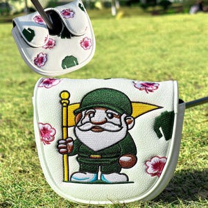 May include: A white golf putter headcover featuring an embroidered gnome wearing a green uniform and holding a yellow flag. The cover is decorated with small pink flower blossoms and green leaf patterns, with a matching smaller cover piece above.