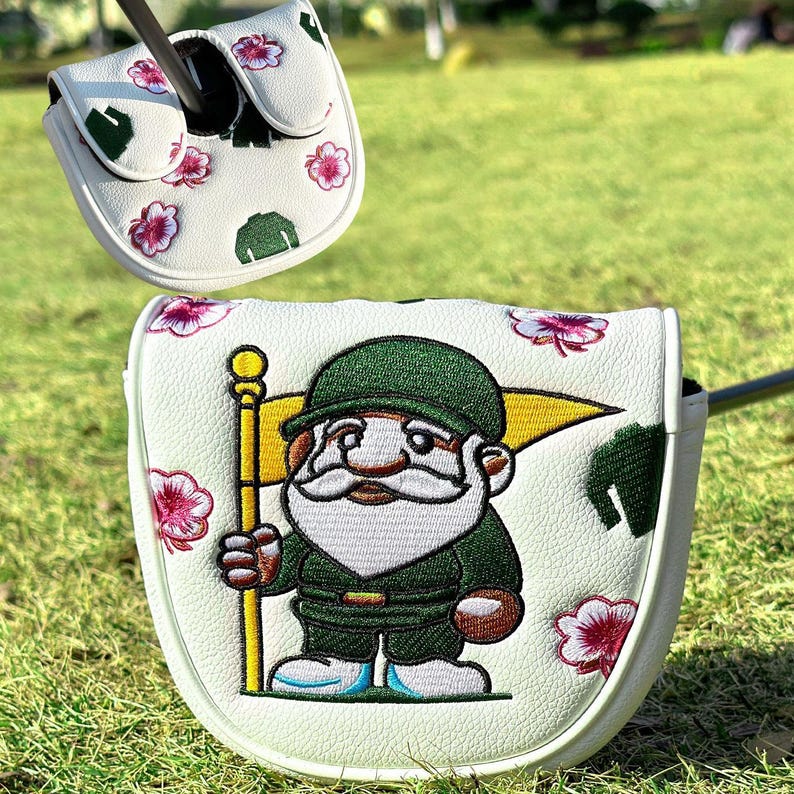 May include: A white golf putter headcover featuring an embroidered gnome wearing a green uniform and holding a yellow flag. The cover is decorated with small pink flower blossoms and green leaf patterns, with a matching smaller cover piece above.