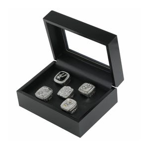 May include: A black wooden display box with a clear glass lid containing five silver-toned championship rings. Each ring features intricate metal detailing, embedded clear stones, and the word CHAMPIONS engraved on the side of the metal bands.