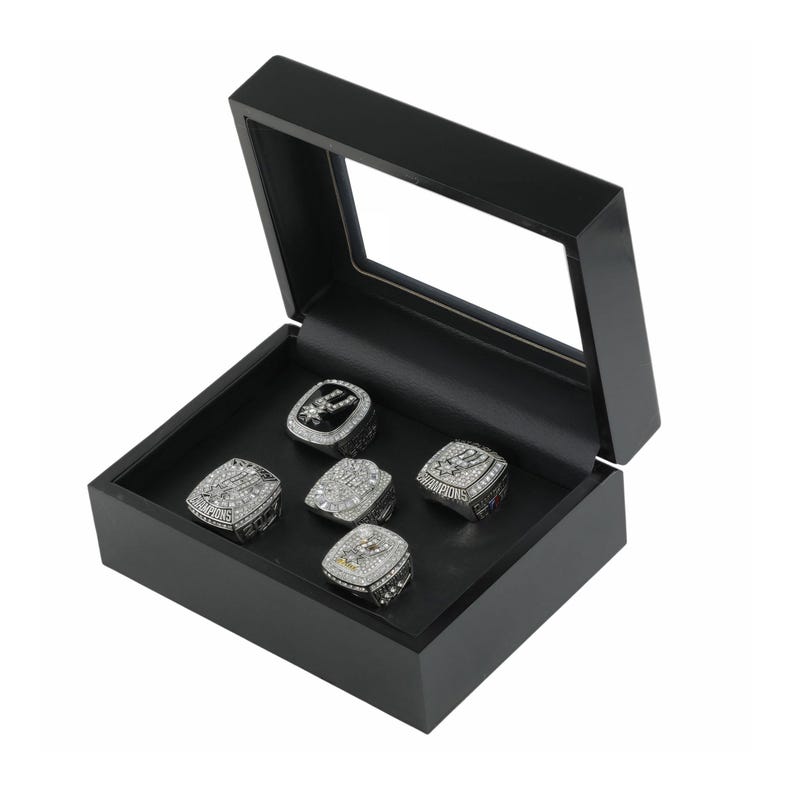May include: A black wooden display box with a clear glass lid containing five silver-toned championship rings. Each ring features intricate metal detailing, embedded clear stones, and the word CHAMPIONS engraved on the side of the metal bands.