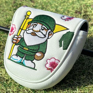 May include: A white golf putter headcover featuring an embroidered gnome wearing a green hat and outfit, holding a yellow flag. The design includes small pink and white floral accents and a green textured fabric detail on a white leather-like surface.