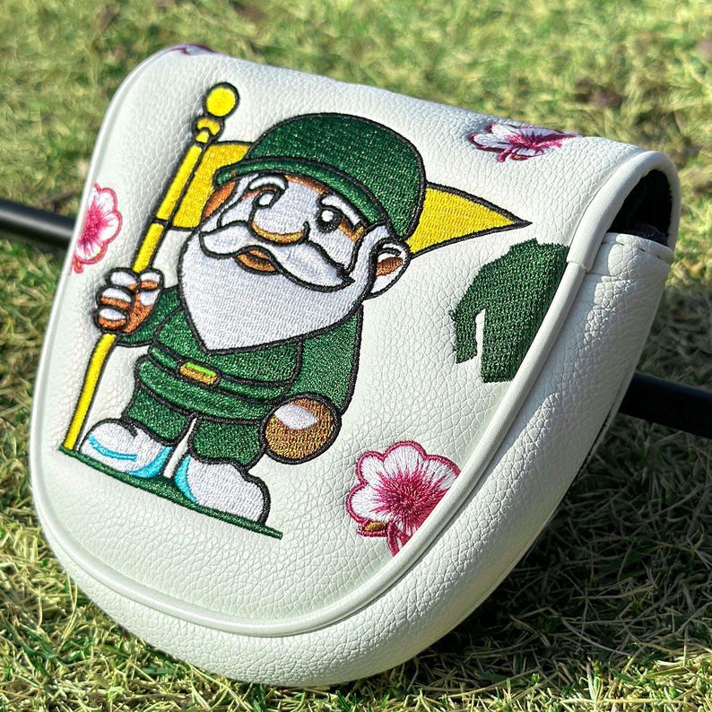 May include: A white golf putter headcover featuring an embroidered gnome wearing a green hat and outfit, holding a yellow flag. The design includes small pink and white floral accents and a green textured fabric detail on a white leather-like surface.