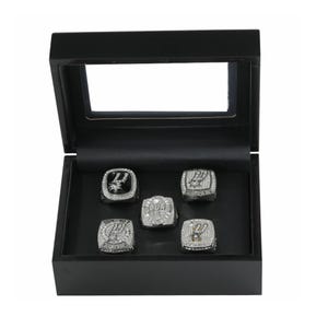 May include: A black wooden display box with a clear lid containing five silver-toned championship rings. Each ring features intricate detailing, embedded clear stones, and the San Antonio Spurs logo. The rings are arranged in a set on a black velvet-lined base.