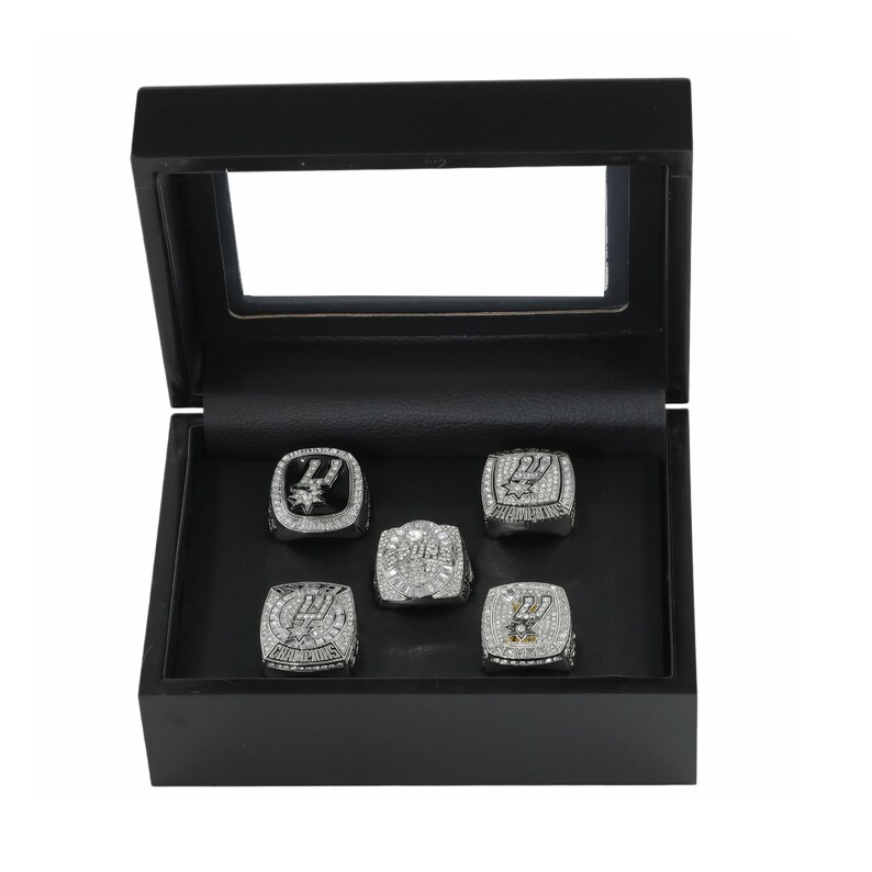 May include: A black wooden display box with a clear lid containing five silver-toned championship rings. Each ring features intricate detailing, embedded clear stones, and the San Antonio Spurs logo. The rings are arranged in a set on a black velvet-lined base.