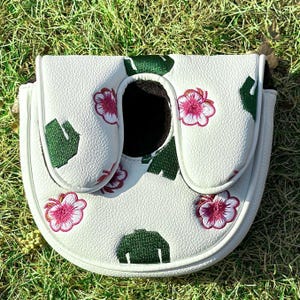 May include: A white mallet-style golf putter headcover featuring a textured faux leather surface. The cover is decorated with embroidered pink hibiscus flowers and dark green sweater icons. It has a central opening and white piping edges.