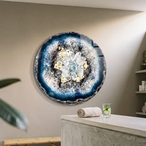 Puede incluir: Una pieza de arte mural circular con un diseño inspirado en una geoda, con anillos concéntricos en tonos azul profundo, blanco y gris. El centro está lleno de racimos de estructuras cristalinas transparentes y acentos dorados, creando una textura.