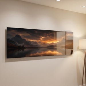 May include: A panoramic wall art print featuring a dark, dramatic mountain landscape at sunset. The scene depicts jagged peaks reflecting in a calm lake under a cloudy, golden-orange sky, with deep shadows and silhouettes of trees along the shore.