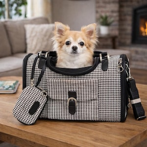 May include: A small, light brown and white Chihuahua dog peeks out of a black and white houndstooth patterned pet carrier. The carrier has black leather accents, a matching pouch, and a shoulder strap. The carrier sits on a wooden table.