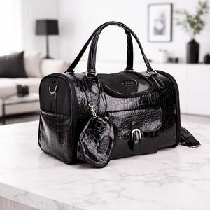 May include: A black pet carrier bag with a glossy, crocodile-skin texture. The bag has a top handle, a side pocket, and a small matching pouch. A silver buckle secures the front pocket. The bag is designed for pet travel.