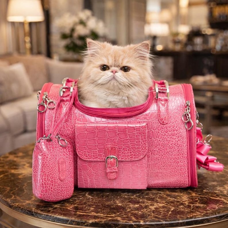 May include: A fluffy, light-colored cat peeks out of a bright pink pet carrier. The carrier has a textured, crocodile-like pattern, a small matching pouch, and silver-toned hardware. The carrier is on a marble-like surface.