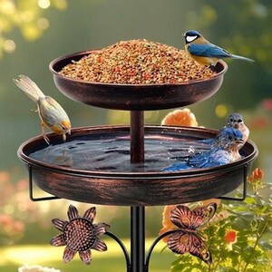 May include: A two-tiered, bronze-coloured bird feeder with a water basin and a food tray. The feeder is decorated with metal flower and butterfly accents. Several birds are perched on the feeder, some bathing in the water and others eating.