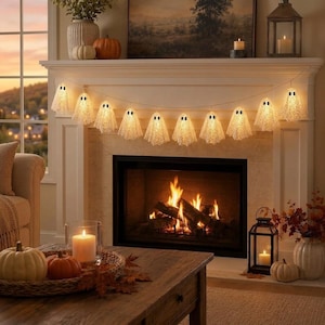 May include: A string of white ghost-shaped lights with black eyes, illuminated and draped across a fireplace mantel. The ghosts are made of a textured, lace-like material, creating a warm glow against the white fireplace and burning fire.
