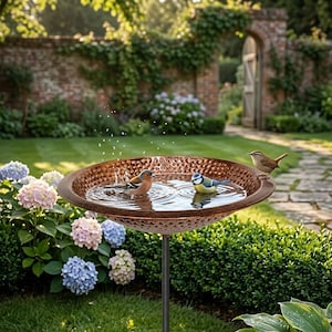 May include: A copper-coloured bird bath with a hammered texture, filled with water. Three small birds are perched on the edge or bathing in the water. The bird bath is on a black metal stand, surrounded by greenery.