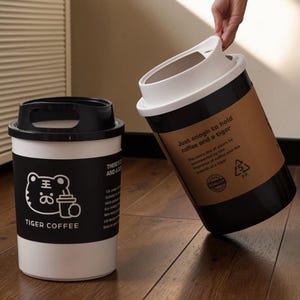 以下が含まれることがあります： 白と黒のコーヒーカップ型の容器が2つ。1つは黒い蓋と、虎のグラフィックと「TIGER COFFEE」の文字が入ったラベルが付いています。もう1つは、「Just enough to hold coffee and a tiger」と書かれた茶色のラベルが付いています。