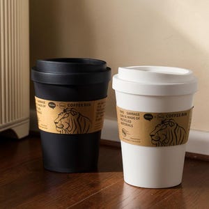 以下が含まれることがあります： 黒と白のコーヒーカップ型のゴミ箱が2つ。それぞれにライオンのロゴと「COFFEE BIN」の文字が入った茶色の紙バンドが付いています。白いゴミ箱のバンドには「THIS GARBAGE CAN IS MADE OF RECYCLED MATERIALS」と書かれています。