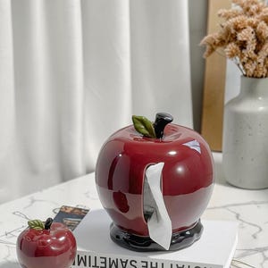 Peut inclure: Un distributeur de mouchoirs en forme de pomme rouge, avec une base noire et une feuille verte. Un petit objet décoratif en forme de pomme est à côté. Le distributeur est posé sur un livre blanc avec le texte "NOT AT THE SAME TIME".
