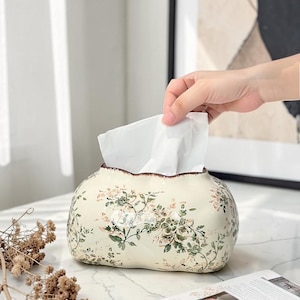 Può includere: Scatola di fazzoletti in ceramica color crema con un motivo floreale rosa e verde. La scatola ha un bordo smerlato ed è piena di fazzoletti bianchi. Una mano si allunga per afferrare un fazzoletto.
