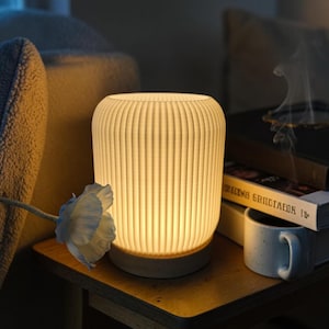 May include: A glowing, off-white ceramic lamp with vertical ribbed detailing. The lamp sits on a small, light-colored base. A white flower and a stack of books are next to the lamp. A mug is also visible.